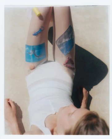 Blonde woman lying on beige fabric, legs up, wearing a white shirt with stickers and toys on her legs in an outdoor setting.