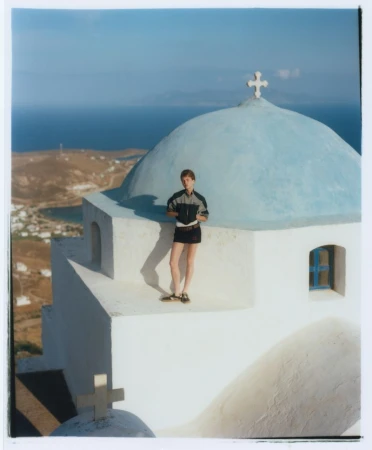 Person stands on Greek island church with blue dome overlooking ocean vista, wearing shorts and jacket in outdoor setting.