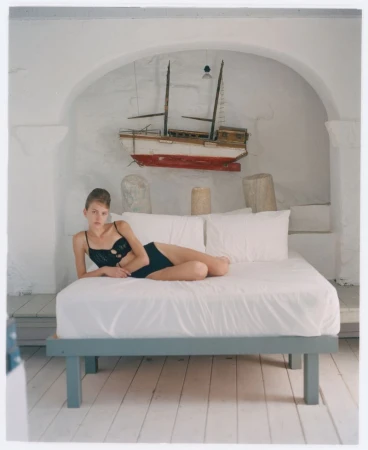 A fair-skinned woman poses in black lingerie on a white bed in a white room with a model boat hanging on the wall above her.