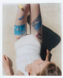 Polaroid photograph of a woman wearing a white swimsuit adorned with small decorative images on her legs, shot from above outdoors.