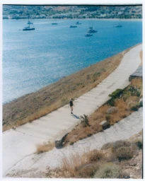 High-angle view of a woman walking down a coastal pathway overlooking a blue sea with anchored sailboats.