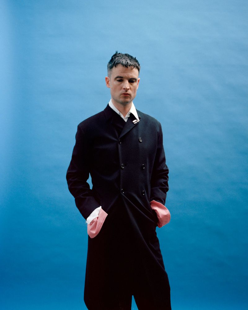 Tom Sturridge wearing a dark buttoned coat with pink cuffs against a blue seamless background for Replica Man Magazine.