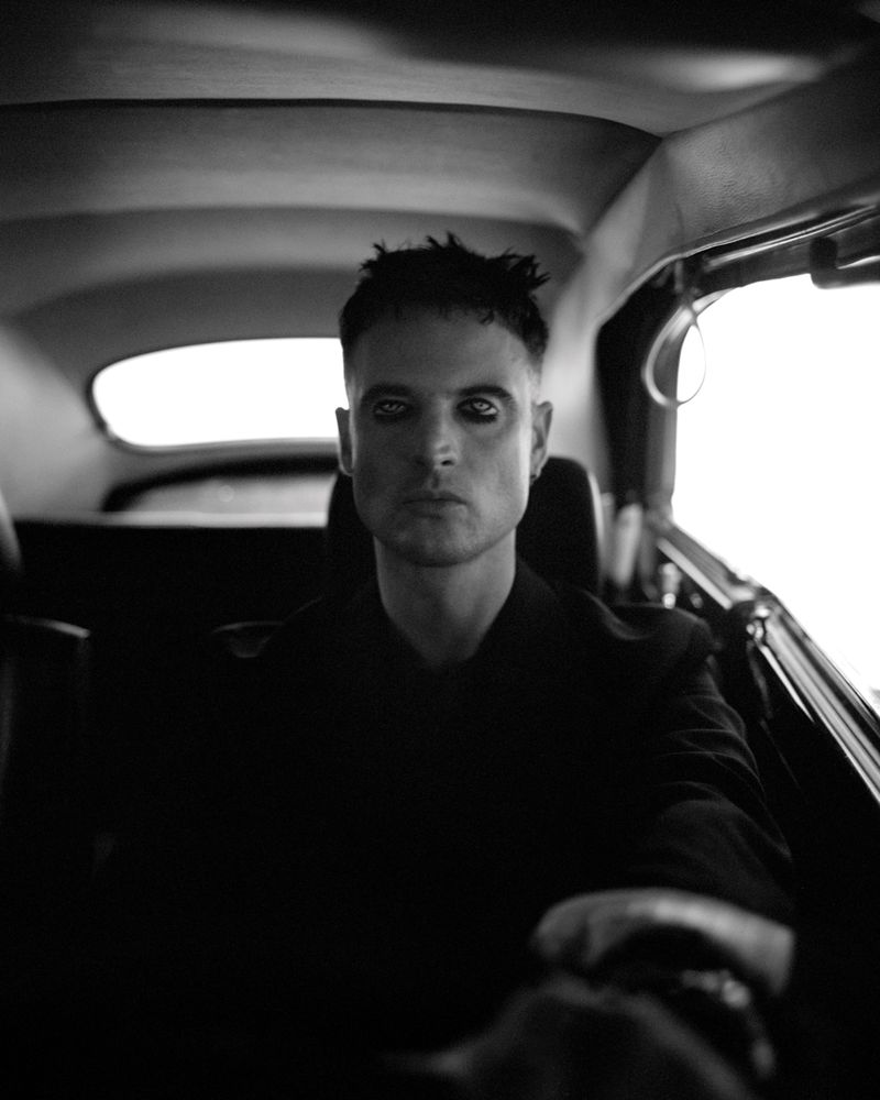 Black and white portrait of actor Tom Sturridge with dark eye makeup sitting inside a vintage car looking directly forward.