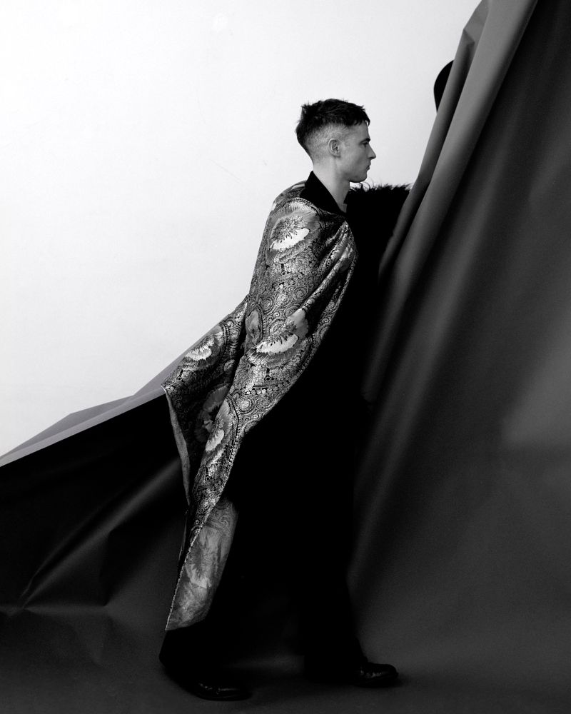 Tom Sturridge wearing an ornate patterned cape over dark clothing, standing beside draped backdrop paper in a studio setting.