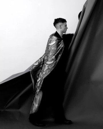Tom Sturridge wearing an ornate patterned cape over dark clothing, standing beside draped backdrop paper in a studio setting.