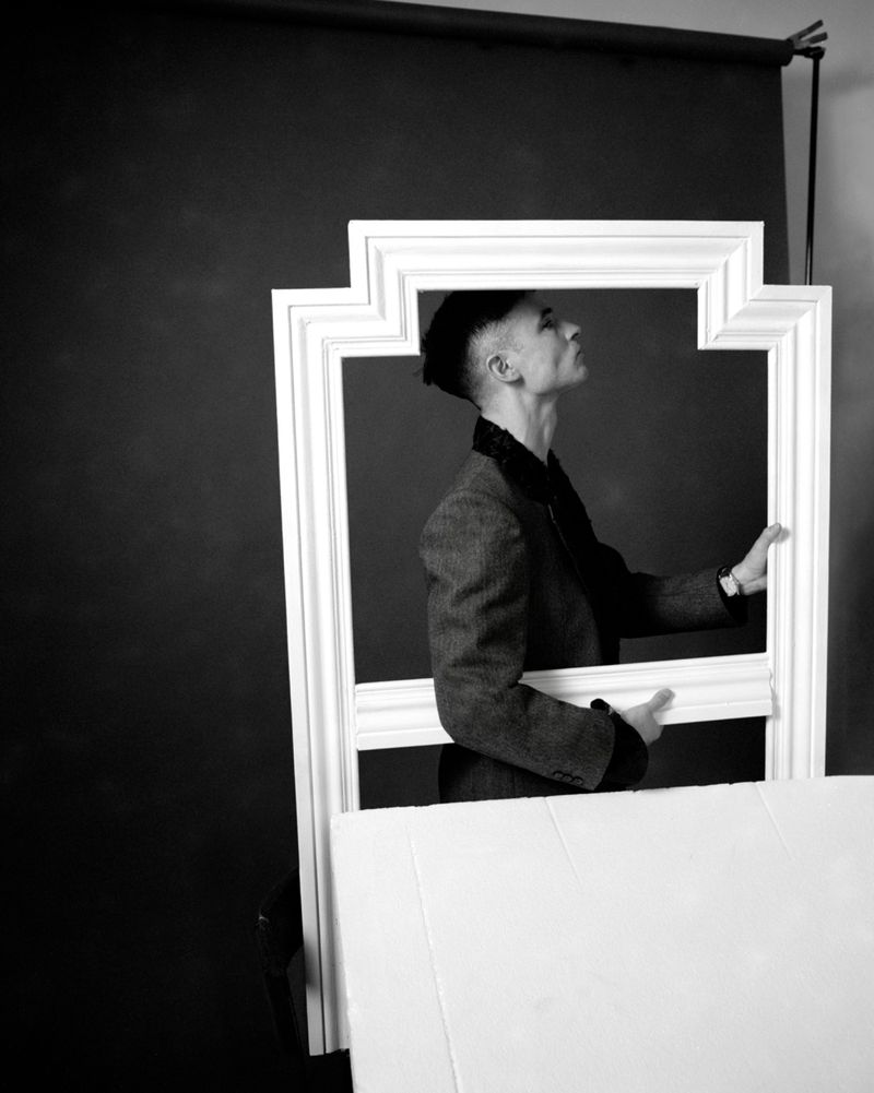 Man wearing a textured jacket looking upward while holding a large white architectural picture frame in a studio setting