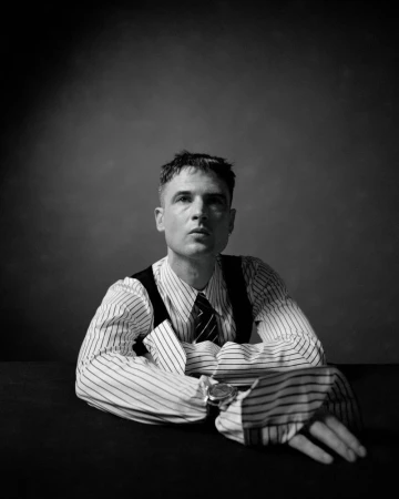 Black and white portrait of Tom Sturridge wearing a striped shirt with oversized cuffs and a tie against a textured studio backdrop.