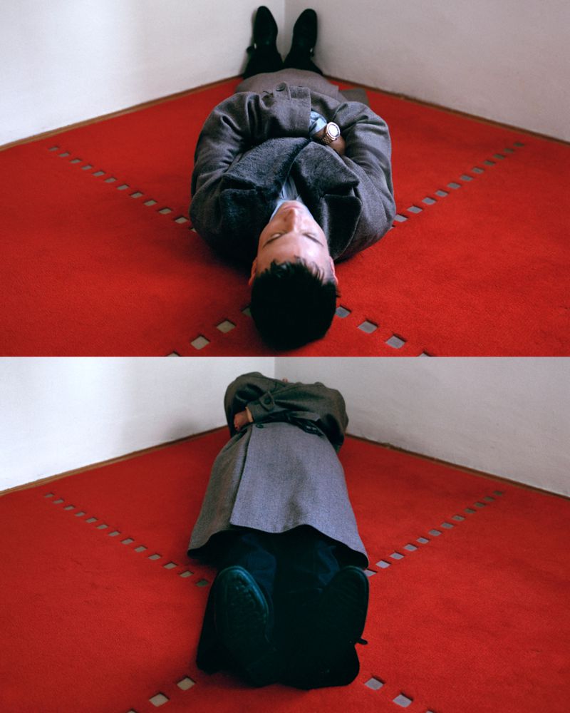 Actor Tom Sturridge lies on a red patterned carpet wearing a grey wool overcoat in a high-angle diptych shot.