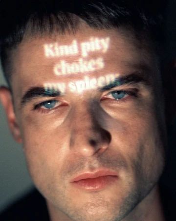 Close-up portrait of Tom Sturridge with glowing text projected across his forehead against a dark background.