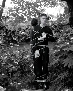 Actor Tom Sturridge tied with rope standing back to back with another figure in an outdoor garden in black and white.