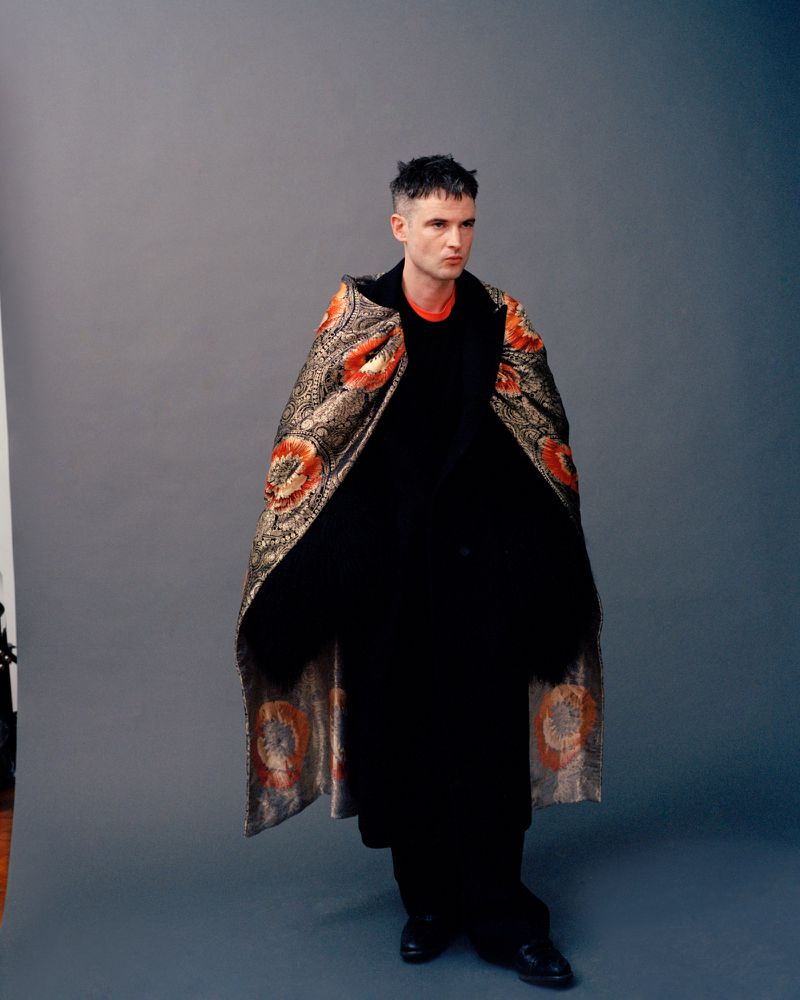 Tom Sturridge draped in a patterned jacquard cape over black attire against a grey studio backdrop.