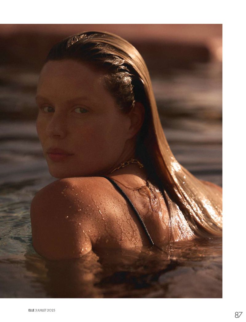 Portrait of a blonde adult female partially submerged in water, wearing a black swimsuit and gold necklace looking over her shoulder.