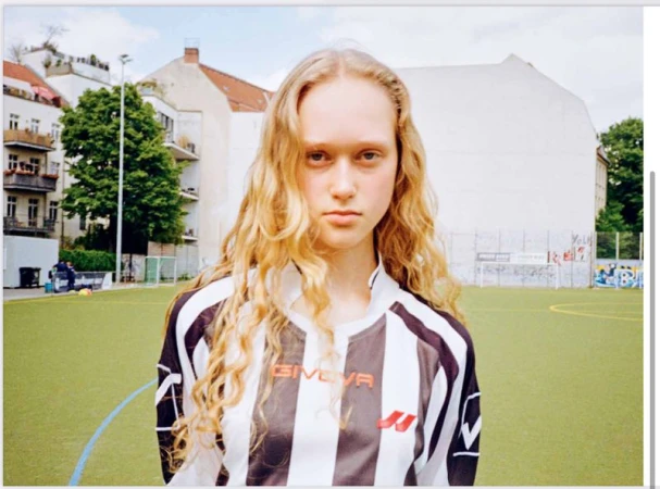 Portrait of a young adult woman with wavy blonde hair, wearing a soccer jersey, standing on a soccer field in an urban environment.