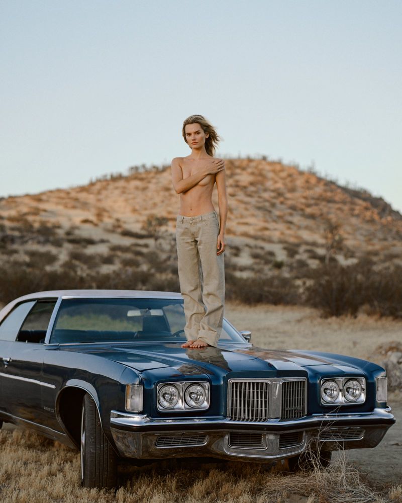 Model standing barefoot on the hood of a vintage dark blue car in a desert landscape for Marie Claire Czech Republic.