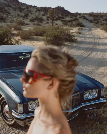 Out of focus woman in red sunglasses standing in front of a vintage blue car parked on a desert dirt road.