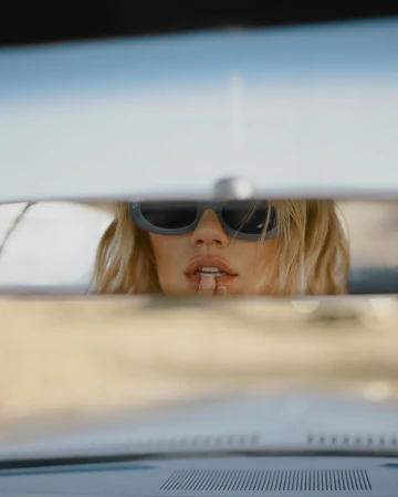 Close-up reflection of a blonde woman wearing oval sunglasses in a car rearview mirror.