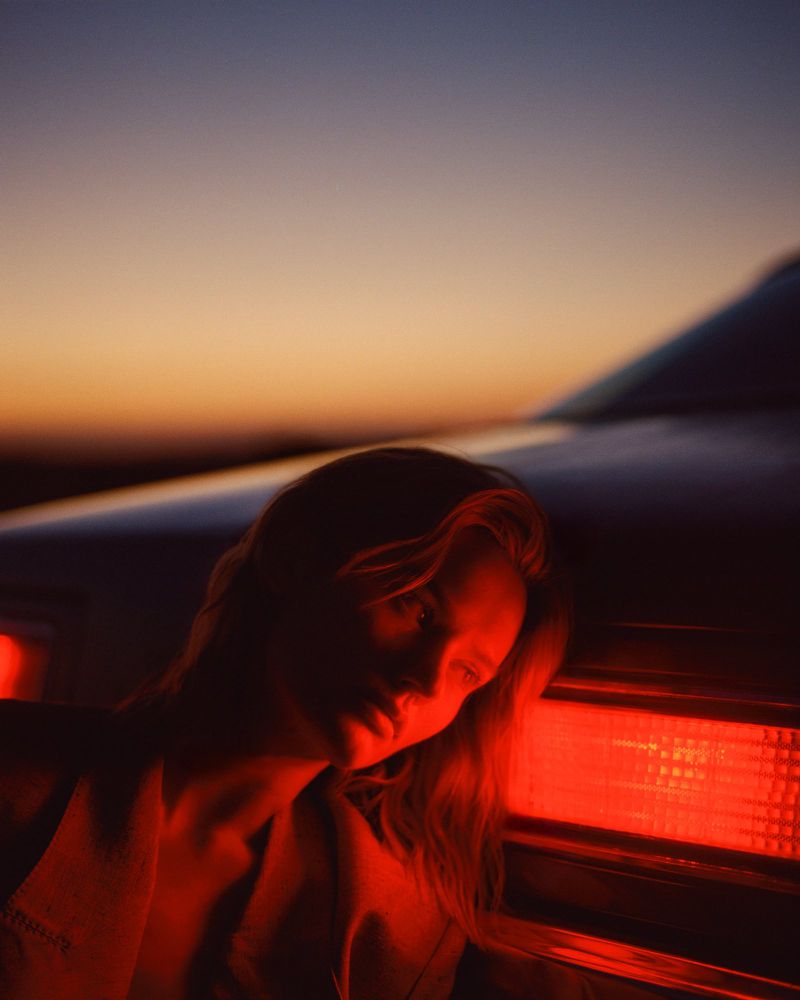 Woman leaning her head against a glowing red car taillight at sunset against a dusk gradient sky.
