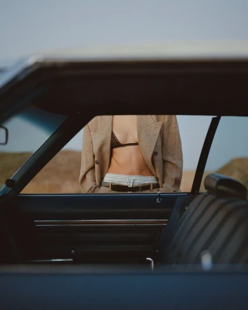 A model wearing an open beige blazer and black bra top framed through a vintage car interior and window.