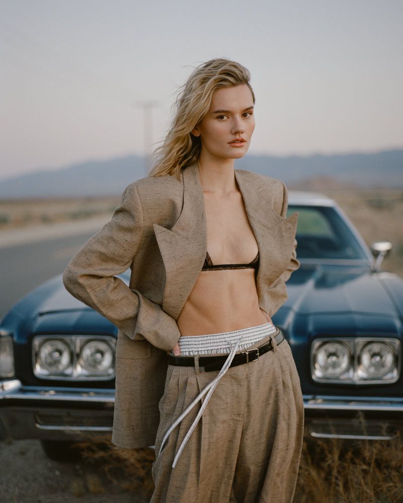 Fashion model posing in an oversized beige suit and black bralette in front of a vintage car on a desert road.