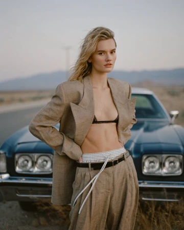 Fashion model posing in an oversized beige suit and black bralette in front of a vintage car on a desert road.