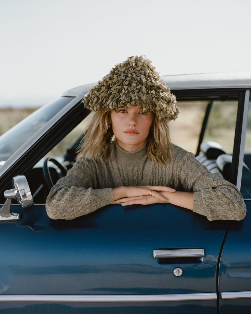 Woman in a textured olive green ruffled hat and knit sweater leaning out of a vintage blue car window.