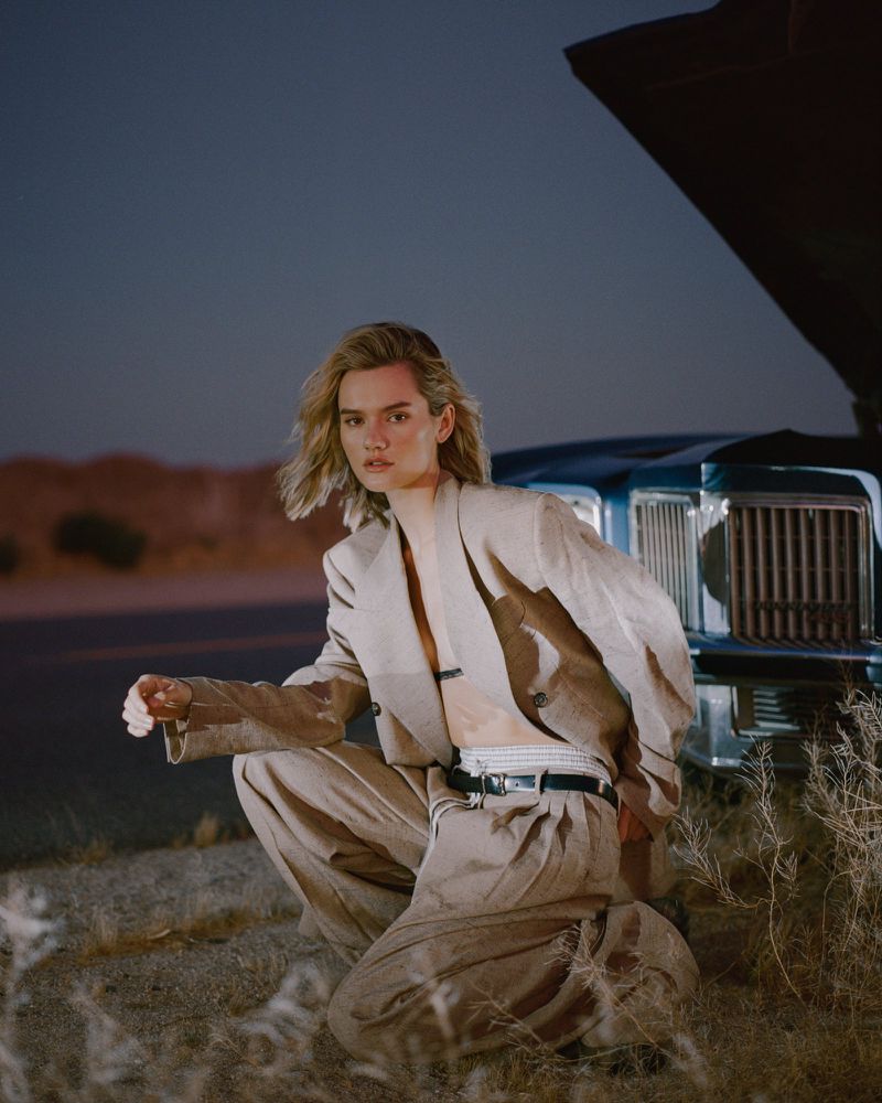 Model crouching beside a vintage car with an open hood on a desert roadside, wearing an oversized beige tailored suit.