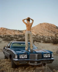A woman stands on the hood of a vintage dark blue car with her back to the camera against a desert mountain landscape.
