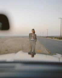 Model wearing an oversized tailored beige suit standing by an open desert highway viewed from inside a car.