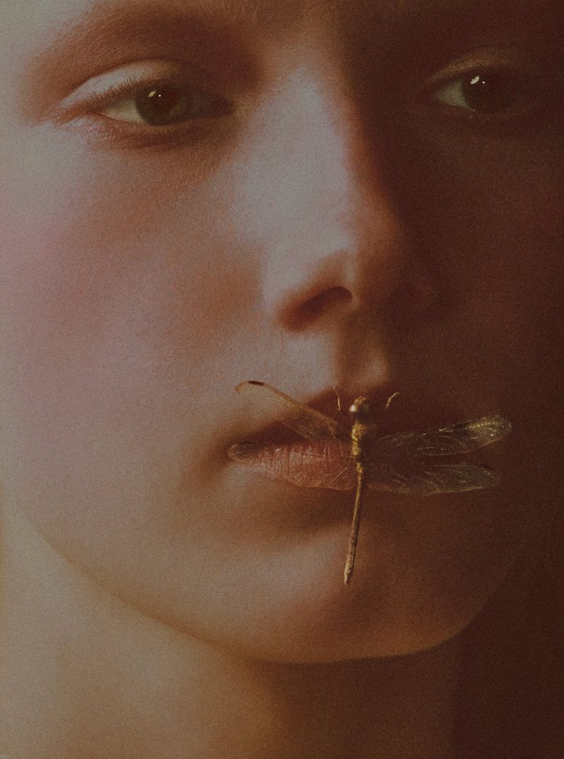 Close-up portrait of model Ivy Stewart with a dragonfly resting across her lips in warm, soft-focus tones.