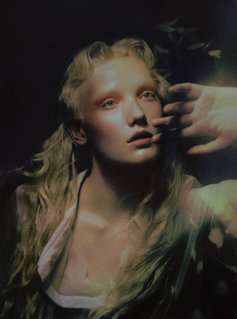 Model Ivy Stewart with blonde wavy hair posing with a raised hand in painterly chiaroscuro light against a dark background.
