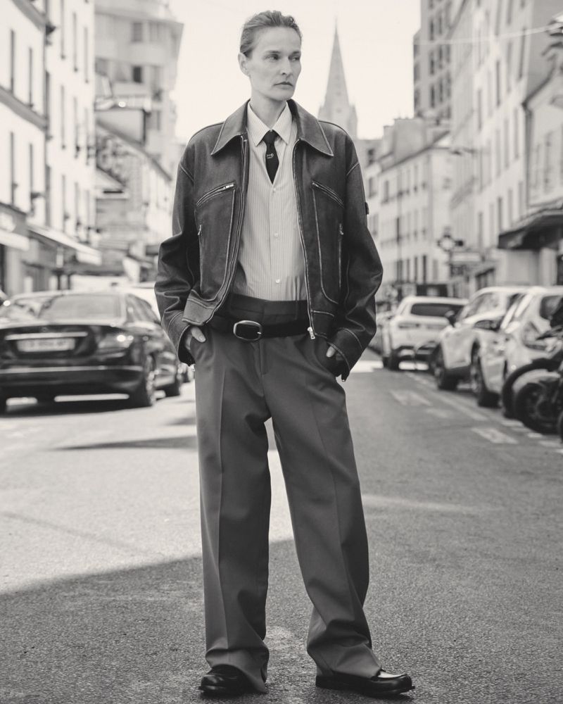 Monochrome full shot of a stylish adult in jacket, shirt, trousers, and shoes on a city street, with buildings and vehicles visible.