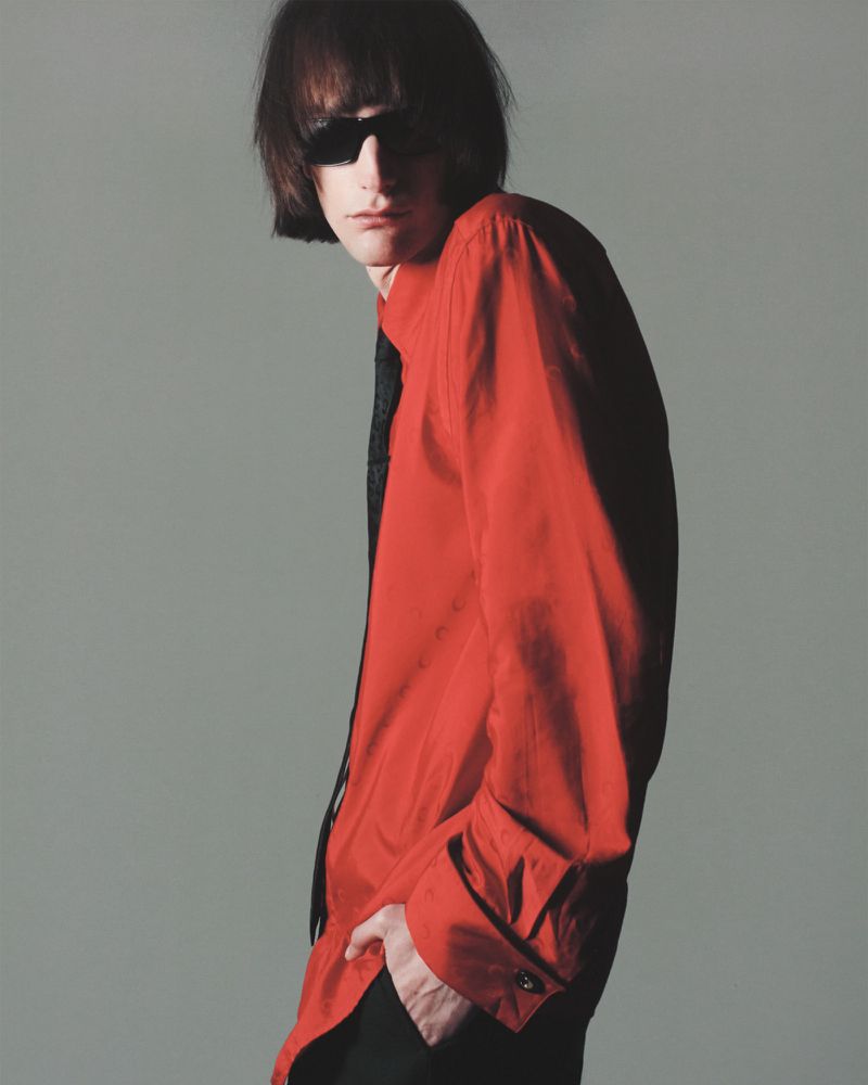 Adult male with dark hair and sunglasses posing in a red button-down shirt with a black tie against a gray background in a studio shot.