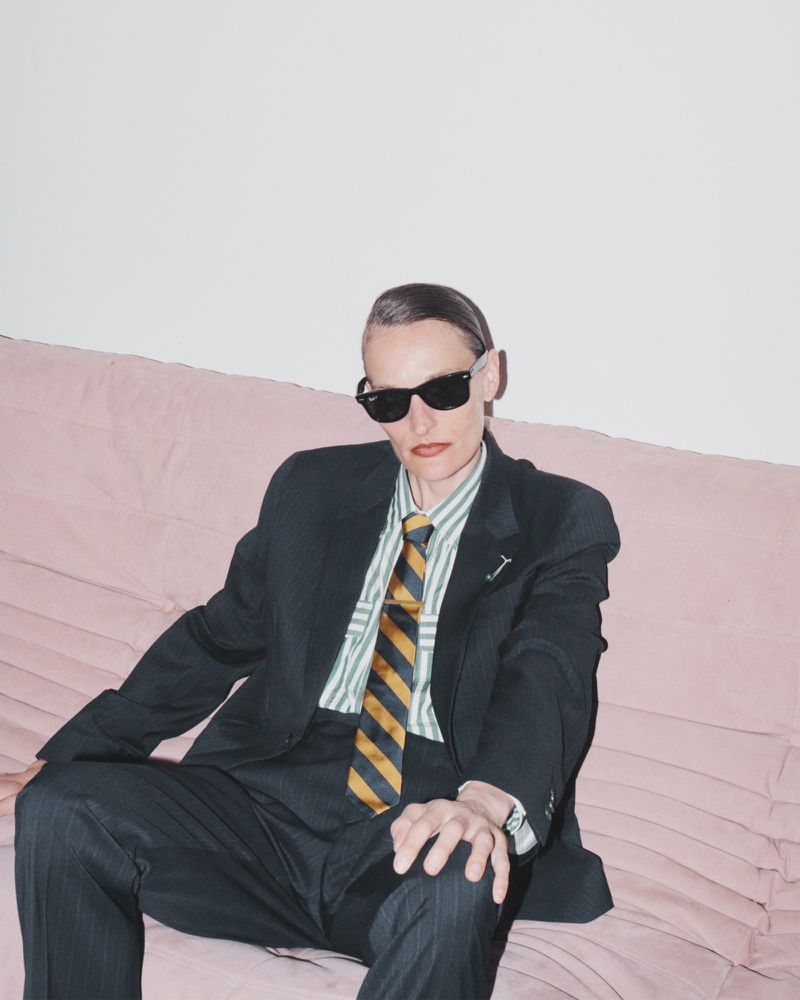 Adult wearing a black suit, sunglasses, and a striped shirt with a tie, seated on a pink couch against a white background.