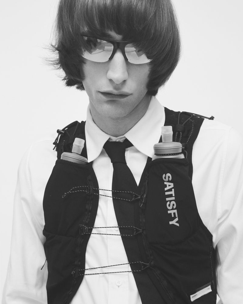 Monochrome portrait of a male fashion model wearing sunglasses, a white dress shirt, a tie, and a black running vest with water bottles.