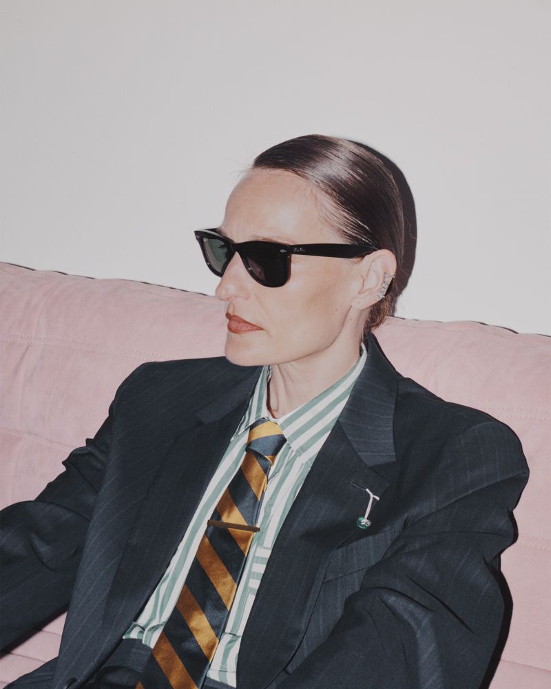 Stylish woman with sunglasses sitting on pink couch wearing dark suit, striped shirt, and tie, in an indoor setting with a white wall.