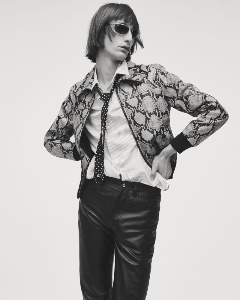 Monochrome fashion portrait of a male model wearing a snakeskin jacket, polka dot tie, and sunglasses against a stark white backdrop.