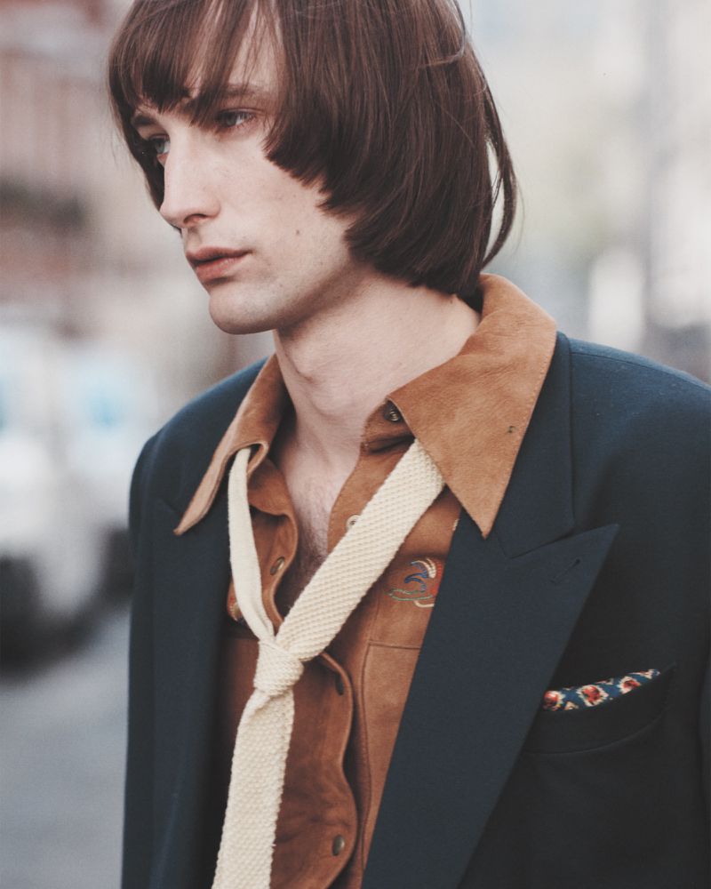 Portrait of a male model wearing a navy blazer, tan shirt and cream tie, standing outdoors on a city street looking downward.