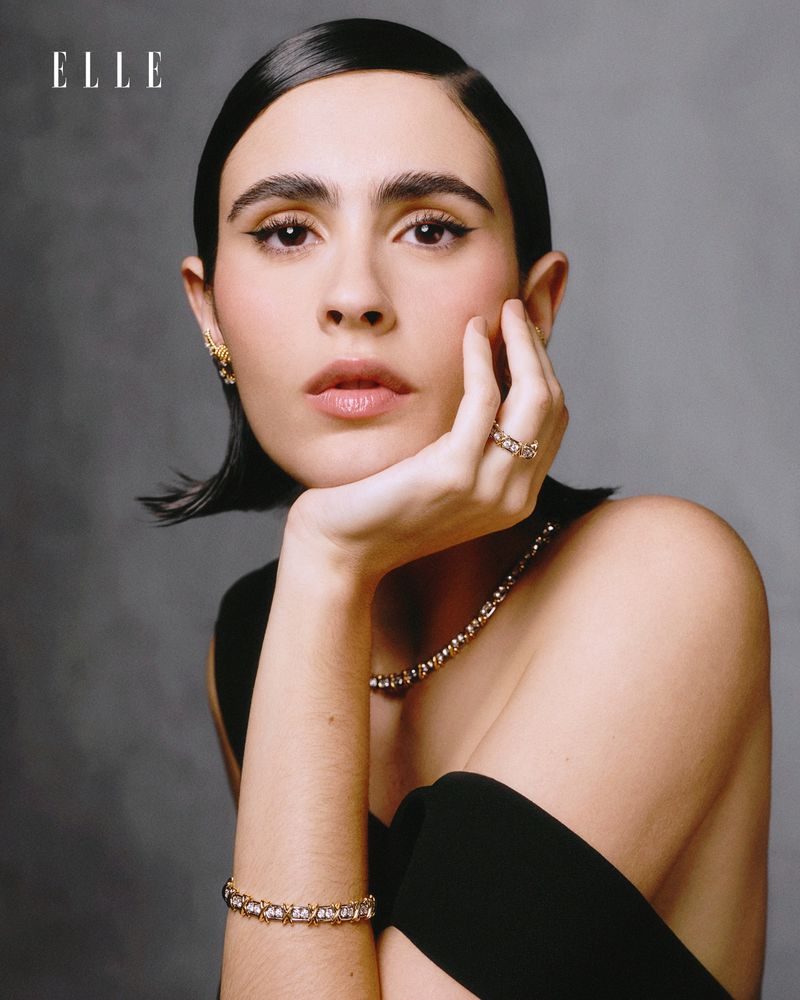 Elegant woman with gold jewelry and a black top poses in a portrait for a magazine cover against a dark grey background.