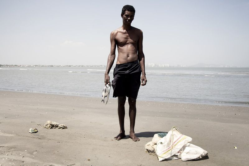 Shirtless man standing barefoot on a sandy beach holding a fish in his right hand with the ocean behind him.