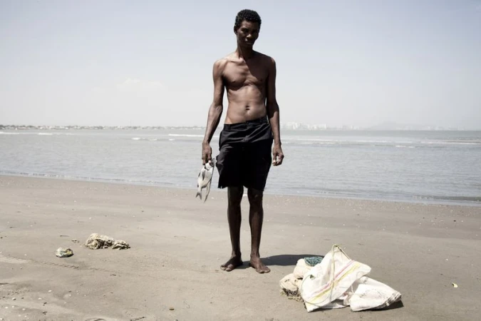 Shirtless man standing barefoot on a sandy beach holding a fish in his right hand with the ocean behind him.