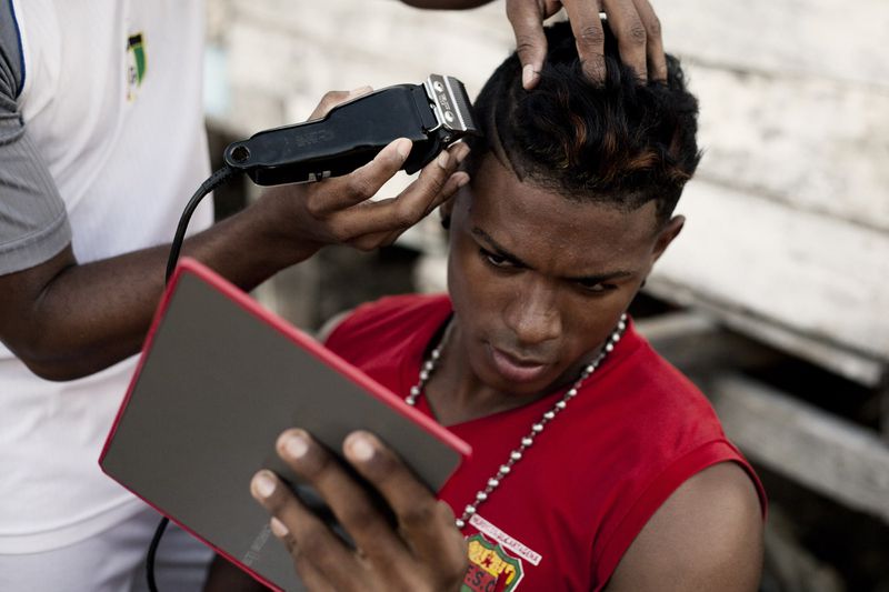 Young Black man in a red sleeveless shirt holding a handheld mirror while receiving a haircut with electric clippers.