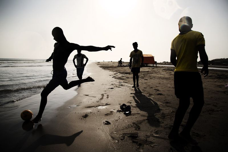 Silhouette of a man kicking a soccer ball on a sandy beach alongside other players under bright sun.