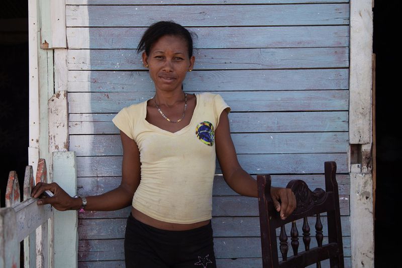 A woman in a yellow shirt stands against a weathered blue wooden wall, resting her hands on a fence and wooden chair.