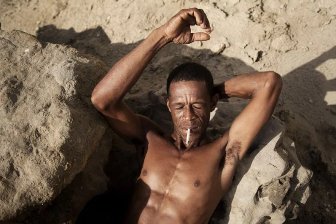 A shirtless man resting back against sunlit rocks with eyes closed and a cigarette in his mouth.