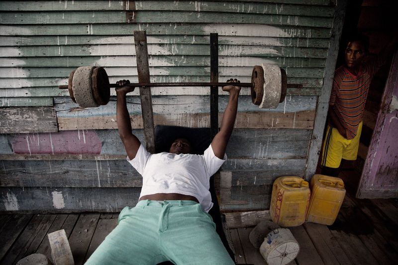 Man bench-pressing a barbell with concrete weights on a wooden bench beside weathered wooden wall planks.