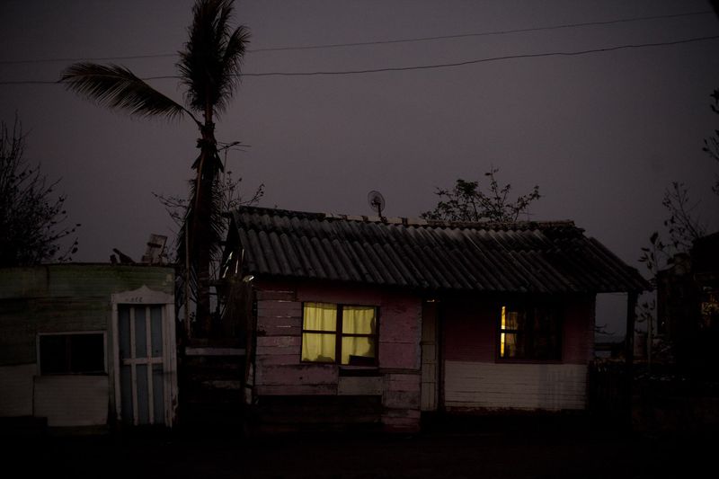 A small rustic house with glowing yellow windows at night under a dark overcast sky beside a tall palm tree.
