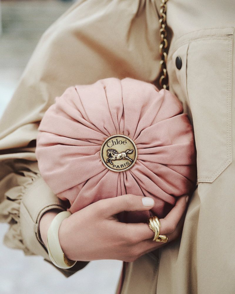 A close-up of a model holding a round pink gathered Chloé pouch bag with a gold horse medallion against a beige trench coat.