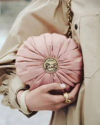 A close-up of a model holding a round pink gathered Chloé pouch bag with a gold horse medallion against a beige trench coat.