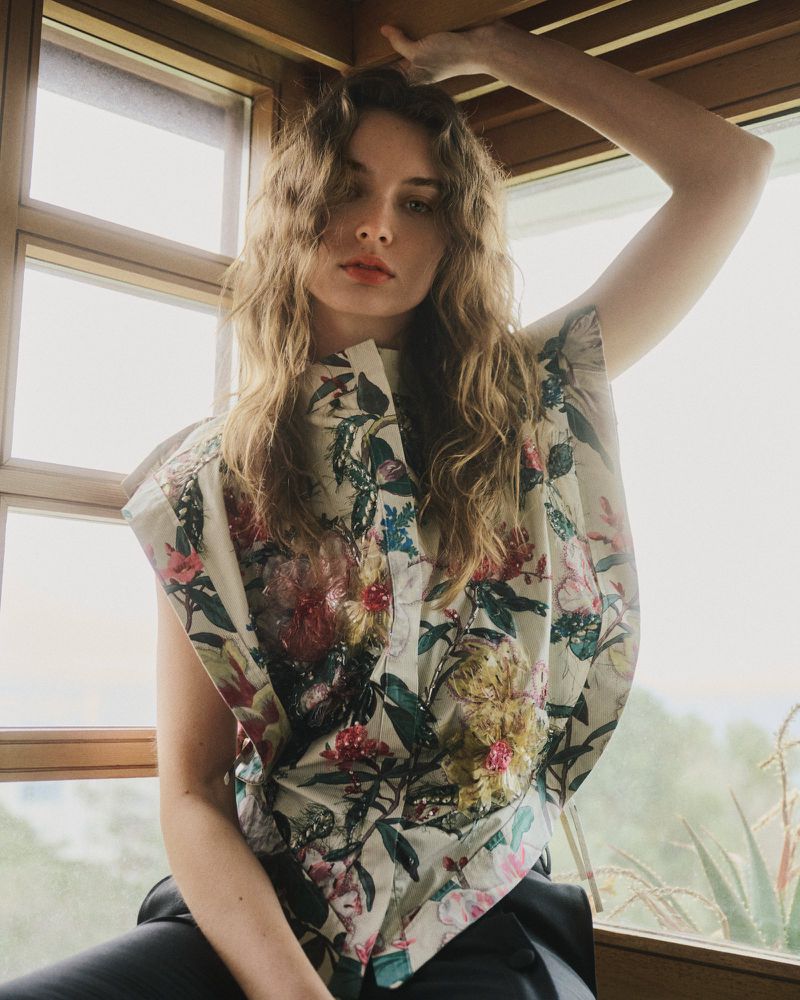 Model Giulia Maenza wearing a floral print sleeveless top seated beside a wood-framed window.