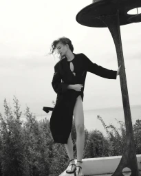 Model Giulia Maenza wearing a black slit coat and strap sandals, leaning against a dark metal structure outdoors.